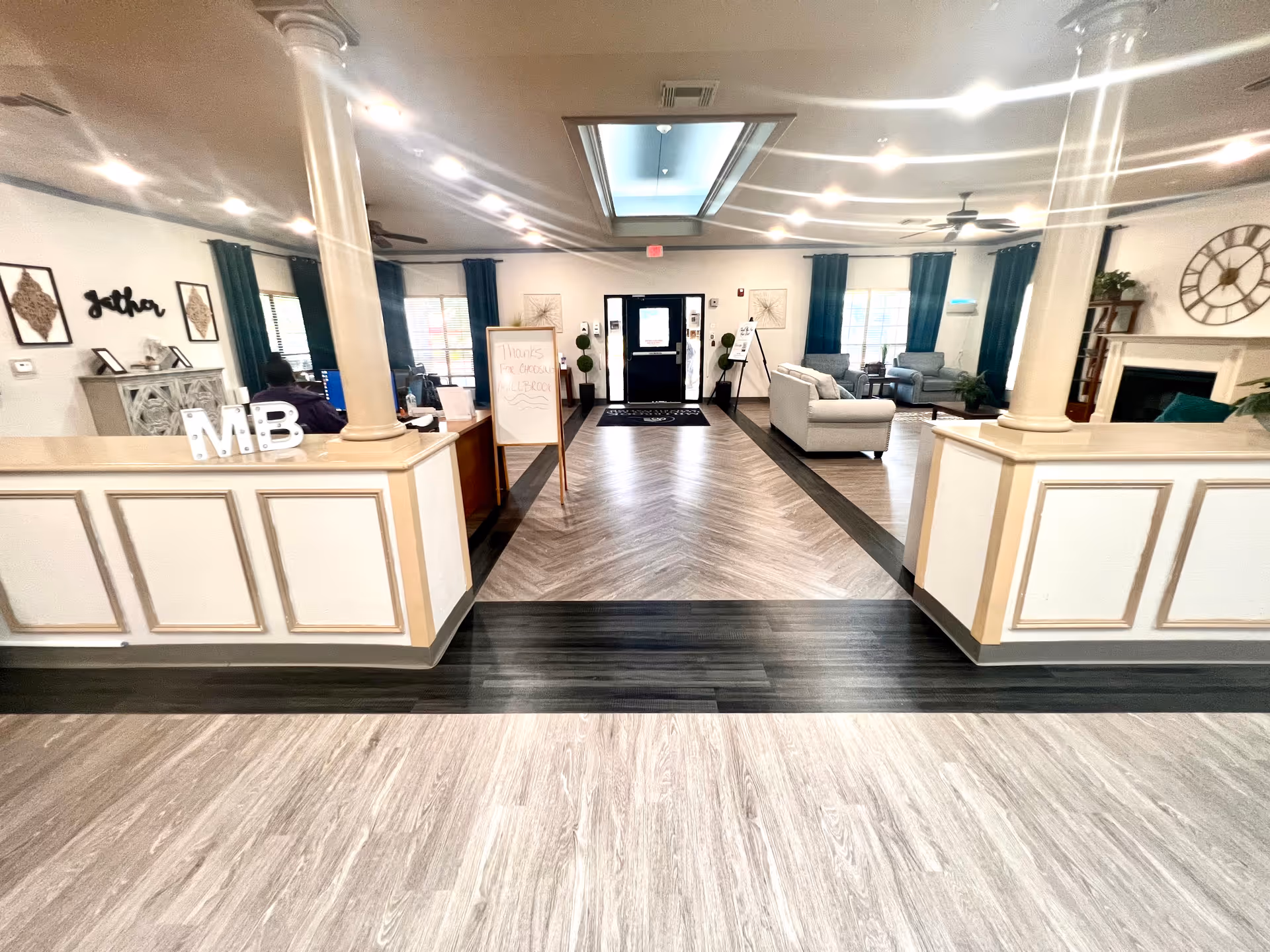 Interior view of a senior living facility lobby or common area with a reception desk on the left, seating area with sofas and chairs on the right, decorative columns, wall art, and a fireplace. The floor has a patterned design and there is a skylight above the central walkway leading to a door at the back.