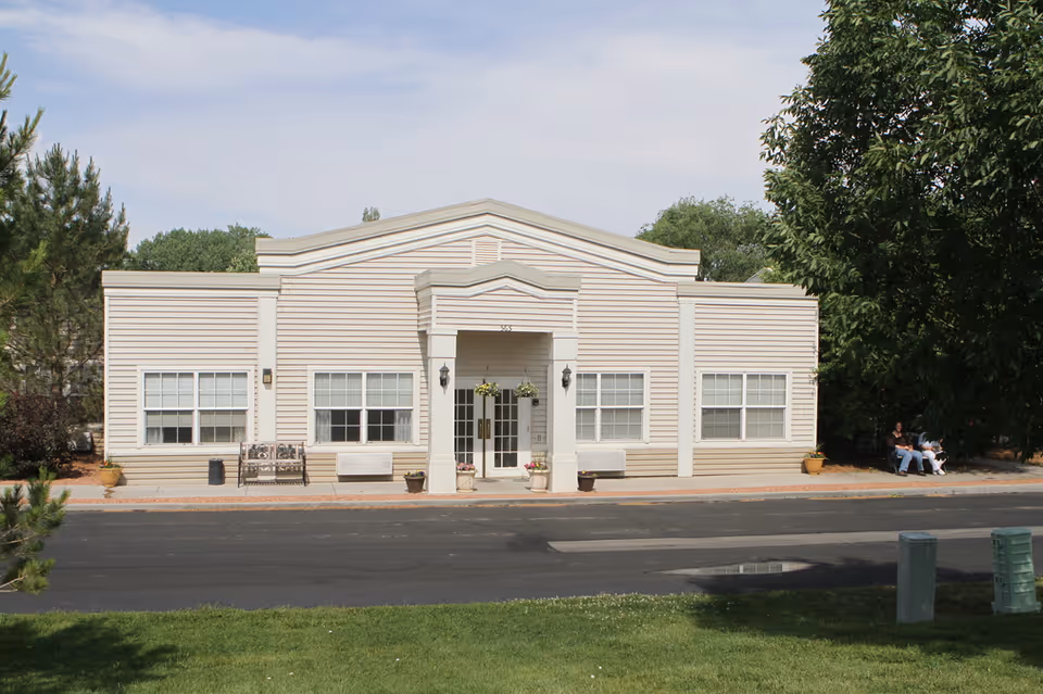 Single-story beige community building with a central covered entrance, windows, benches and potted plants.