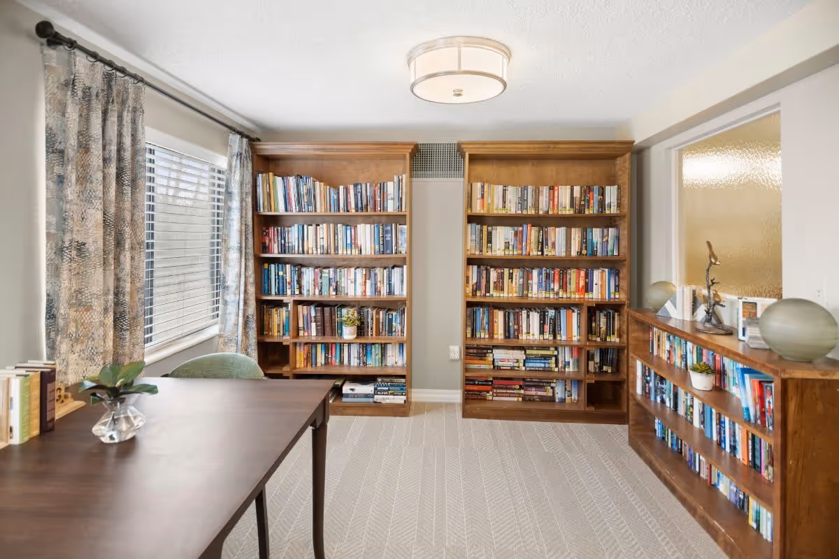 A bright room with two large wooden bookshelves filled with books against the back wall and a lower wooden bookshelf on the right side also filled with books. A wooden table with a small plant in a glass vase and a few books is positioned on the left side near a window with patterned curtains. The room has light-colored walls, a patterned carpet, and a ceiling light fixture.