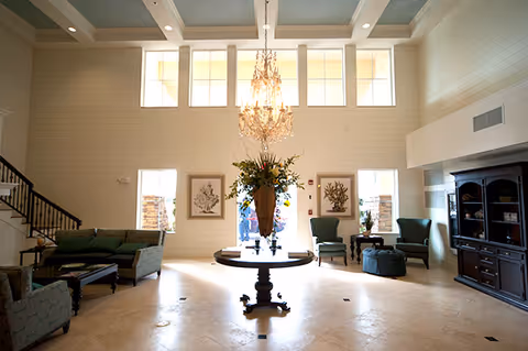 Spacious and well-lit living room area with high ceilings and large windows. The room features a central round table with a large floral arrangement, a chandelier hanging above, green upholstered sofas and armchairs arranged around the space, a dark wooden cabinet on the right, and a staircase on the left.