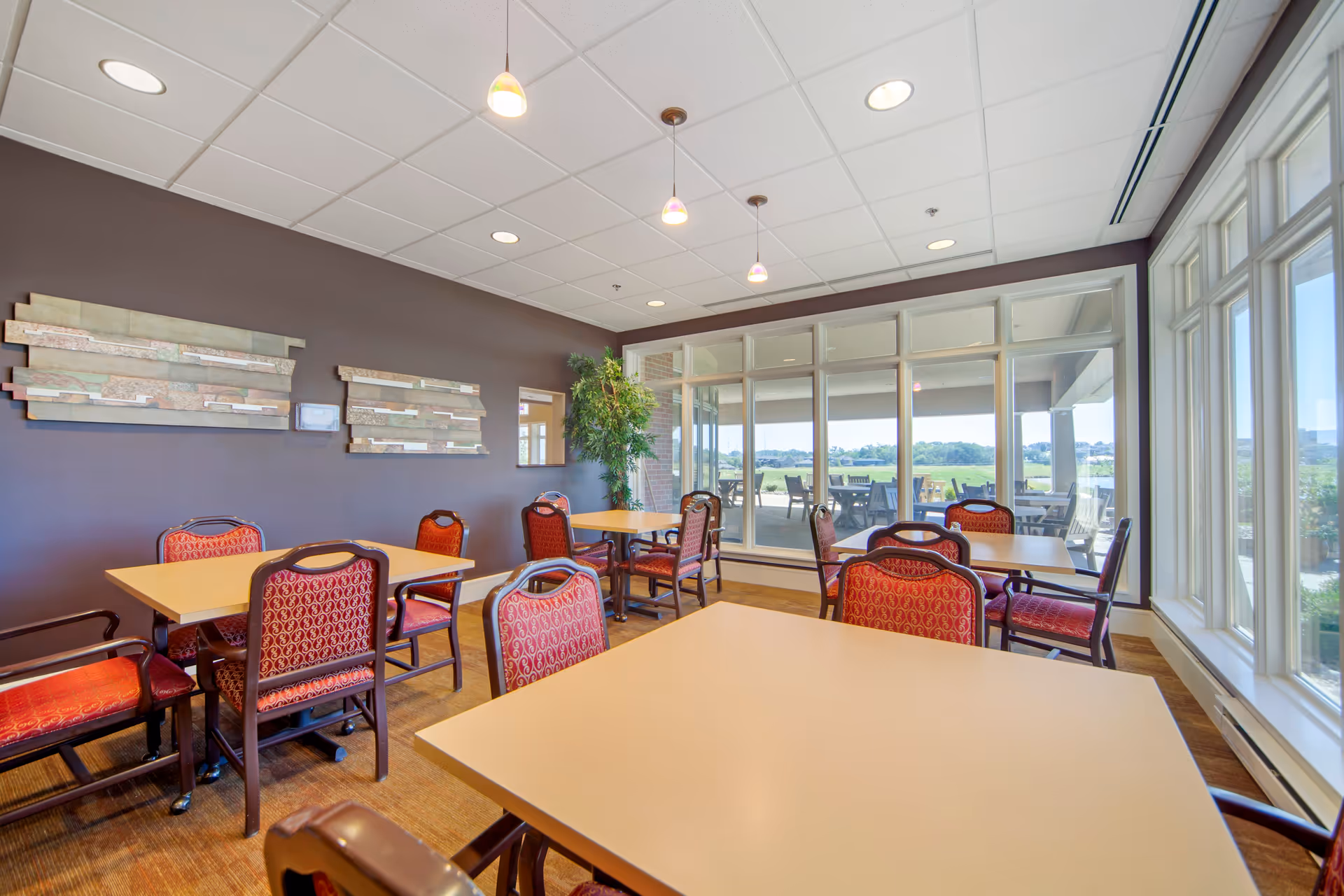 A bright dining room with several tables and red cushioned chairs arranged neatly. Large windows on one side provide a view of an outdoor patio area with additional seating and a green landscape beyond. The walls are painted dark brown with decorative wall art and a potted plant in the corner. Ceiling lights hang above the tables.