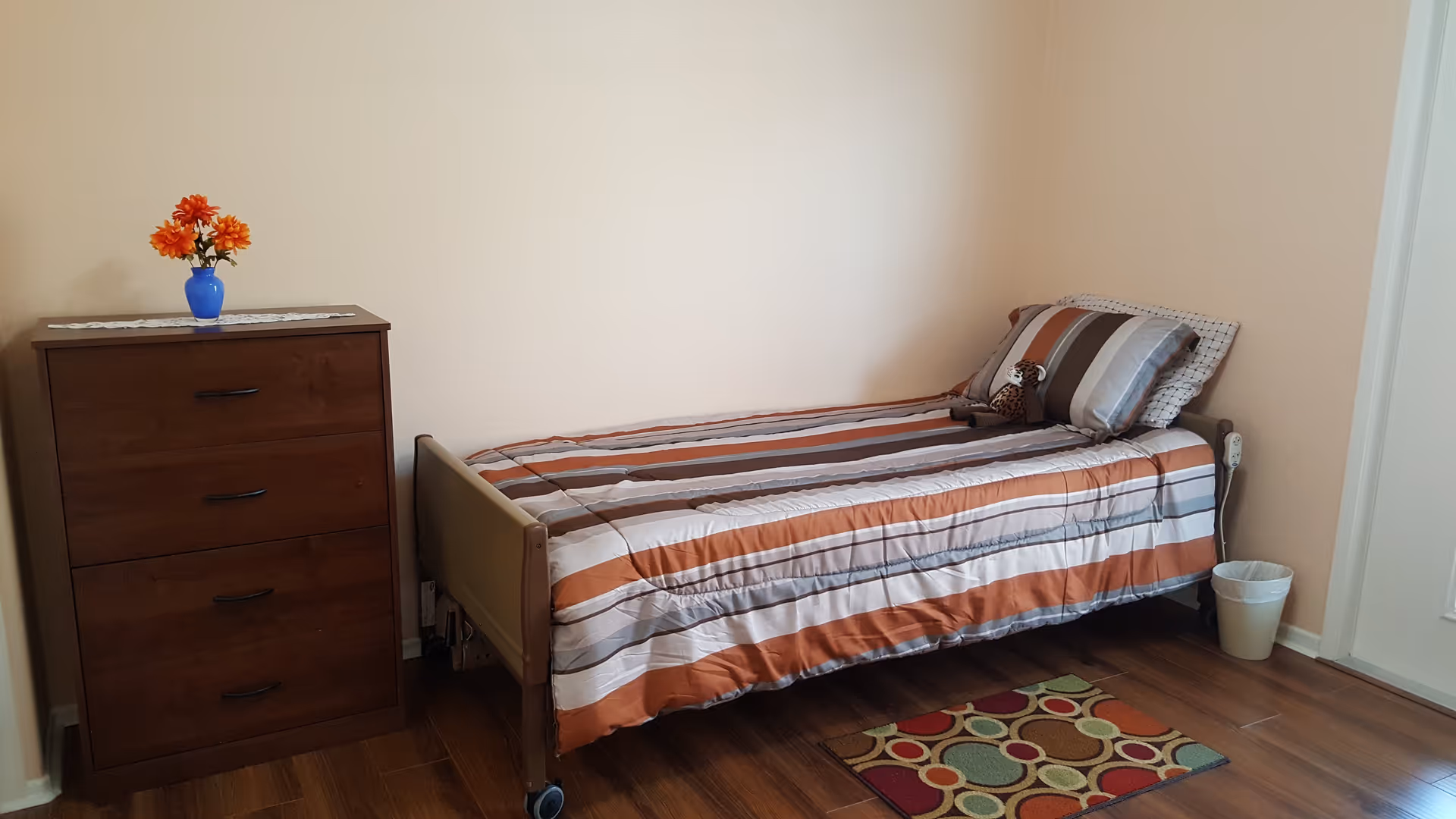 A simple bedroom with a single bed covered in a striped orange, brown, and white comforter. There is a small stuffed animal on the bed. Next to the bed is a wooden chest of drawers with a blue vase holding orange flowers on top. A small colorful rug is on the wooden floor near the foot of the bed, and a white trash bin is placed near the bed against the wall.