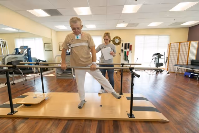An elderly man performs a leg exercise using parallel bars in a rehabilitation room while a female therapist observes and takes notes. The room has wooden floors, exercise equipment, and large windows letting in natural light.