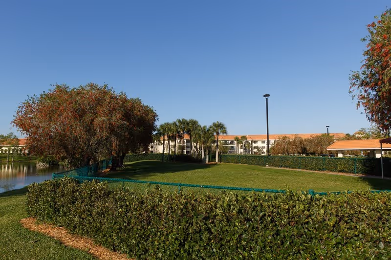 Outdoor view of a senior living facility with a well-maintained green lawn, trees, and a pond on the left side. The building with a red-tiled roof is visible in the background under a clear blue sky.