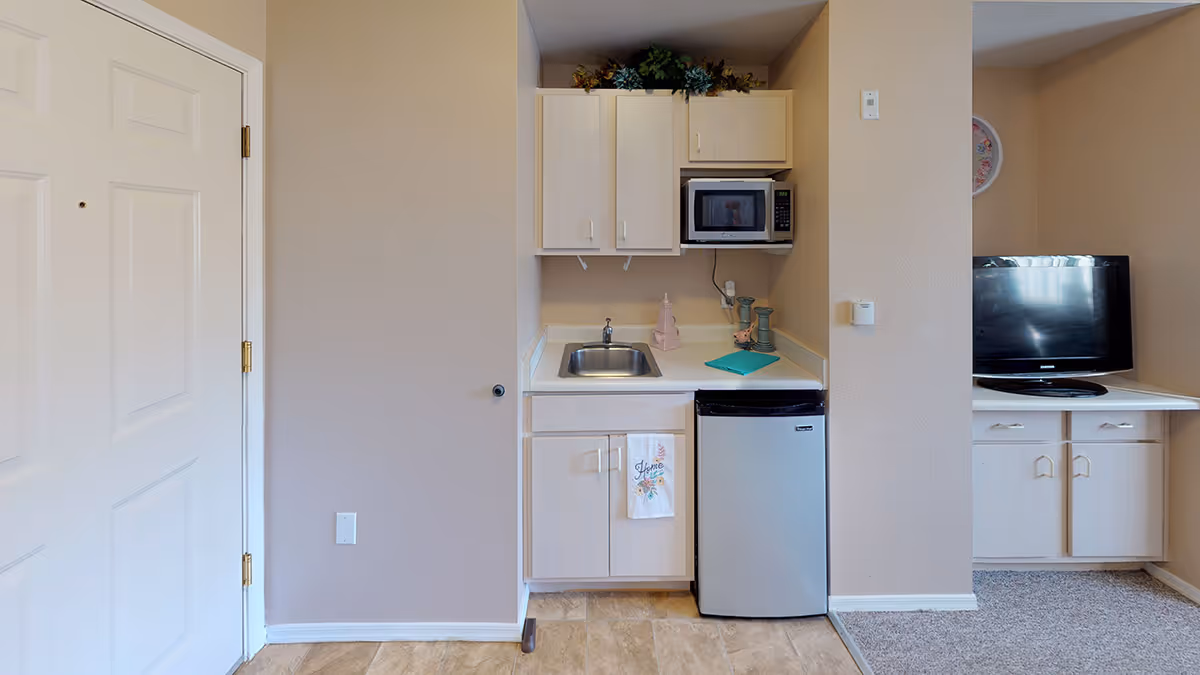 Small kitchenette area with light wood cabinets, a stainless steel sink, a microwave mounted above the counter, a mini refrigerator, and decorative items on the counter and above the cabinets. To the right, a TV sits on a white cabinet in an adjacent living space. The walls are beige and the floor is a mix of tile and carpet.