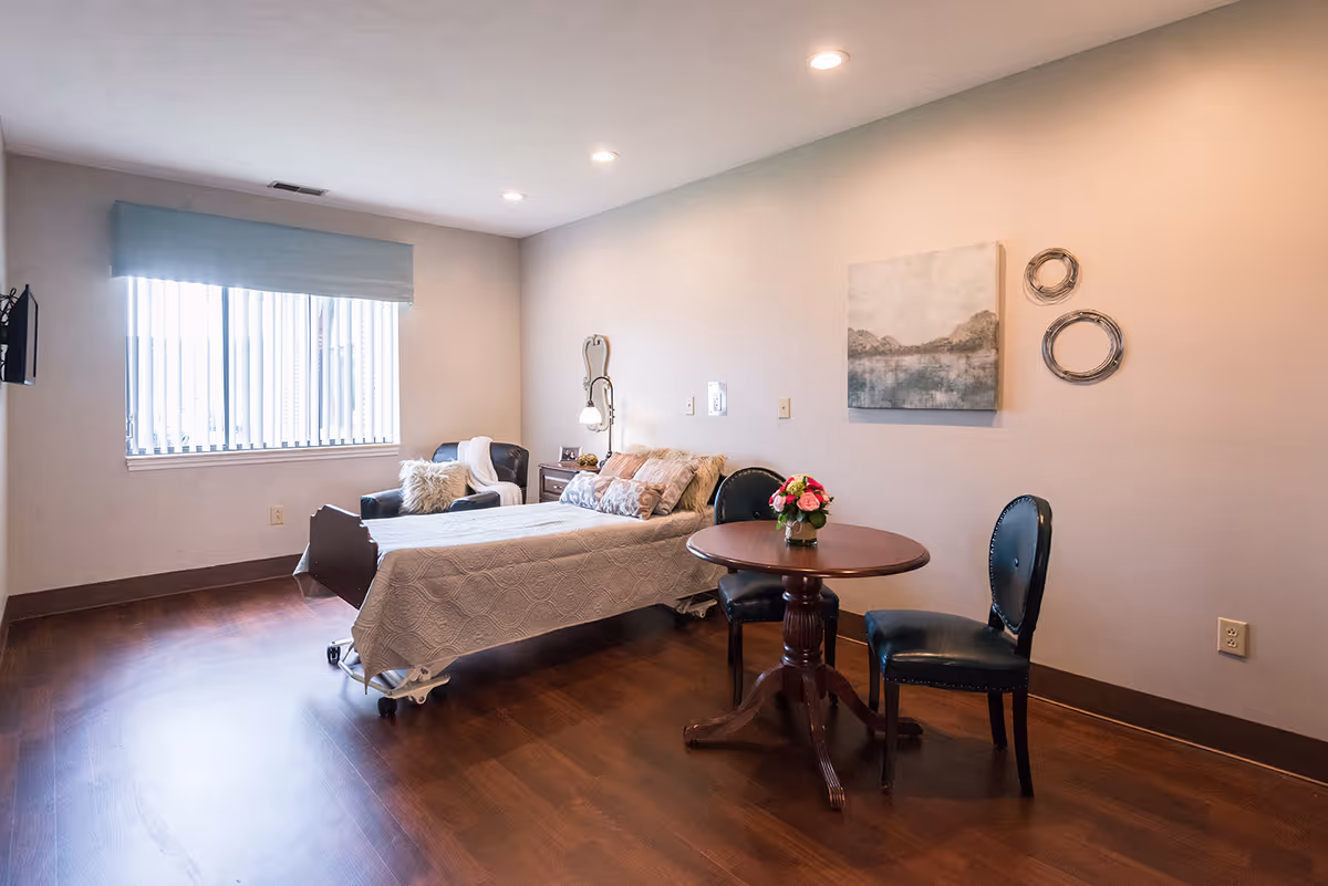 A neatly furnished senior living bedroom with a single bed, a small round table and two chairs, an armchair by the window, and wall decor.