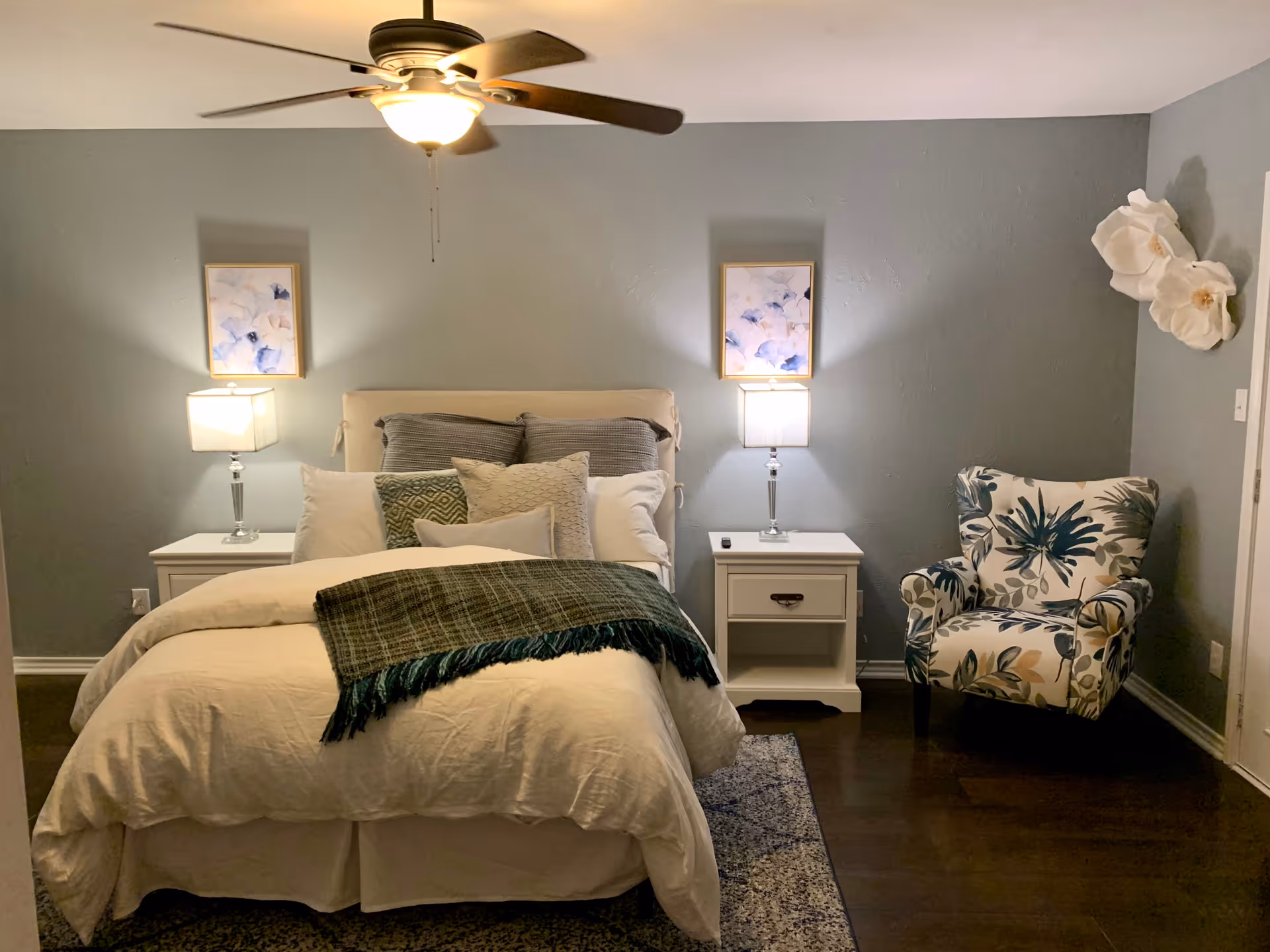 A cozy bedroom with a neatly made bed featuring white bedding and multiple pillows. There are two white nightstands on either side of the bed, each with a lamp and a framed abstract artwork above. A ceiling fan with a light is on the ceiling. To the right of the bed, there is a floral patterned armchair and two large white decorative flowers mounted on the wall. The walls are painted a soft gray color and the floor is dark wood with a patterned rug partially visible under the bed.