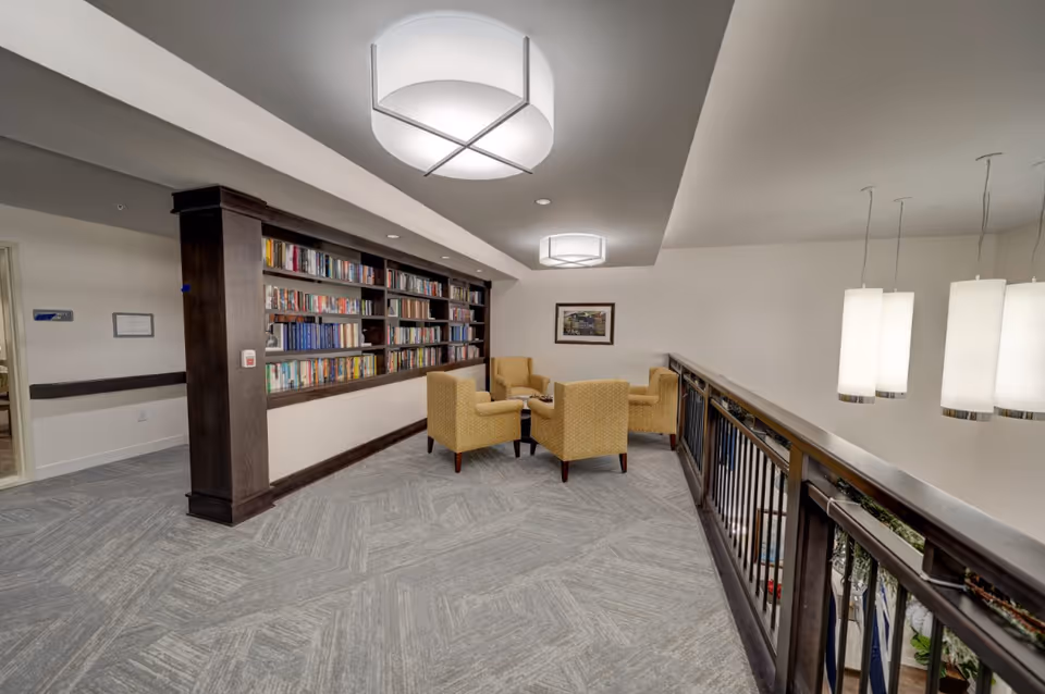 Upstairs library/lounge with built-in bookshelves, a small seating area of four yellow armchairs, and pendant lights over a railing.