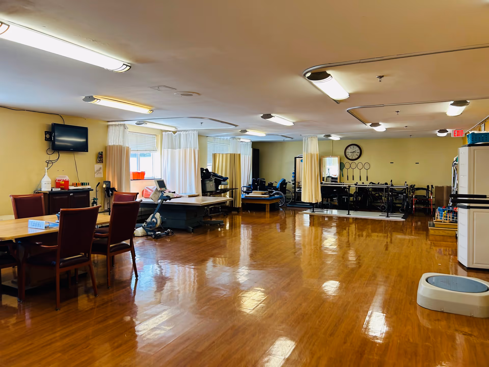 Spacious rehabilitation therapy room with treatment tables, exercise machines, chairs, and polished hardwood floor.