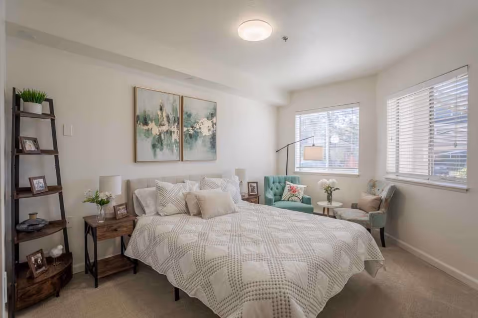 A bright and cozy bedroom with a large bed covered in a white and beige patterned quilt, multiple pillows, and two abstract paintings hanging above the headboard. There is a wooden nightstand with a lamp and flowers on one side, a tall wooden shelf with decorative items on the left, and two upholstered chairs with a small round table and a floor lamp near two windows with white blinds.