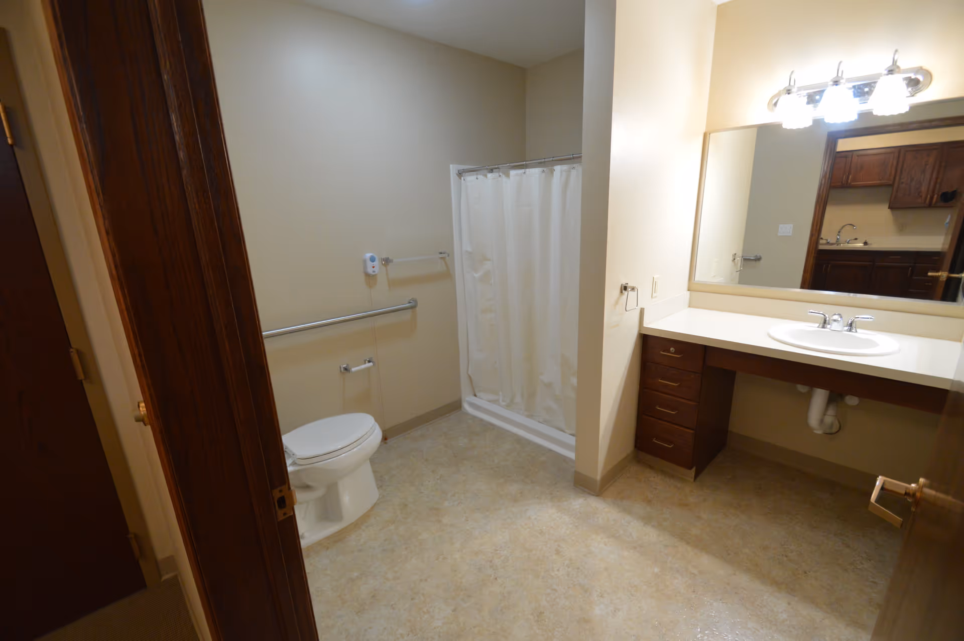 A bathroom with beige walls and flooring, featuring a white toilet with a grab bar beside it, a shower with a white curtain, and a vanity with a sink, countertop, and mirror above. The room has wooden door frames and a light fixture above the mirror.