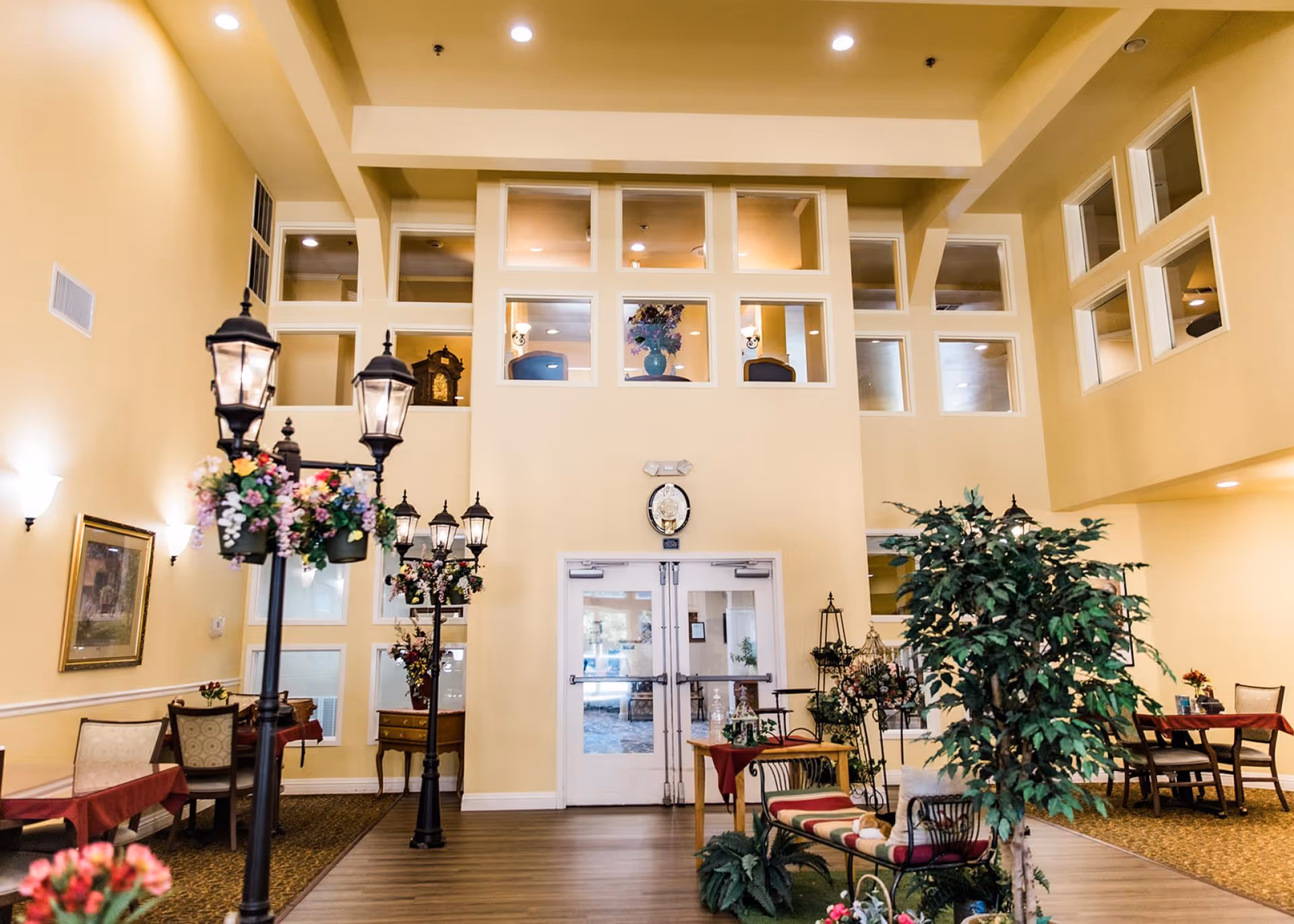 Bright two-story common area with tables, chairs, decorative lamp posts, plants, and a double-door entrance.