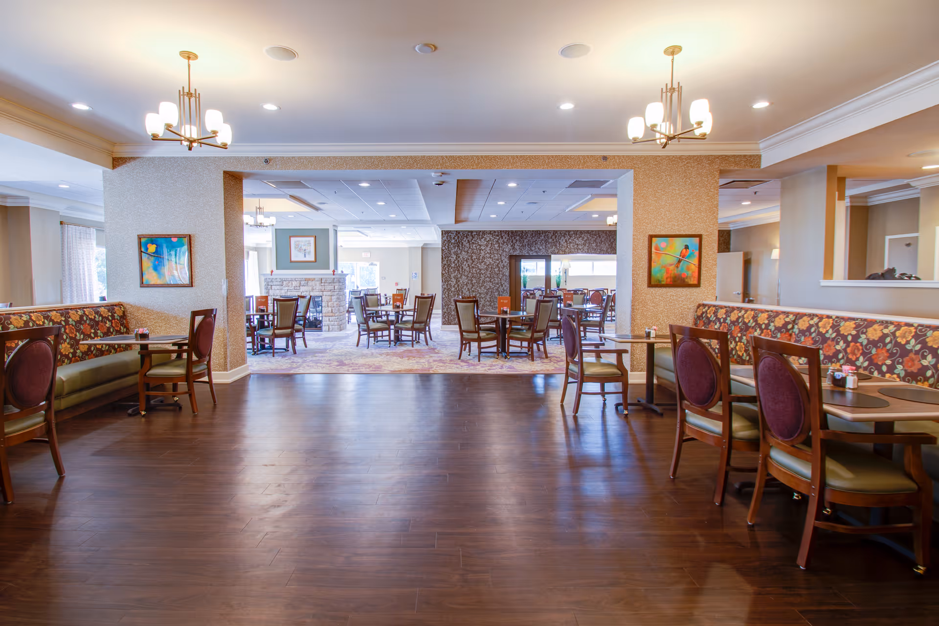 A spacious dining area in a senior living facility with wooden floors, multiple tables and chairs, floral patterned booth seating along the walls, modern chandeliers, and colorful artwork on the walls. The room extends into another dining space with more tables and chairs and a stone fireplace in the background.
