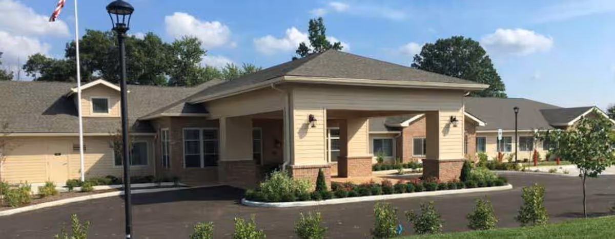 Exterior view of a single-story senior living facility building with a covered entrance, beige siding, brick accents, a flagpole with an American flag, and landscaped greenery under a partly cloudy sky.