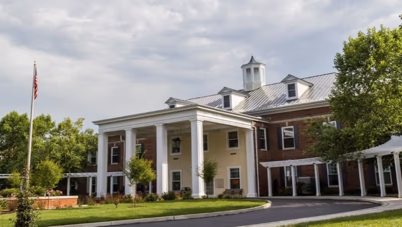 Front exterior view of a large brick building with white columns and a covered entrance, surrounded by green trees and a lawn, under a cloudy sky.