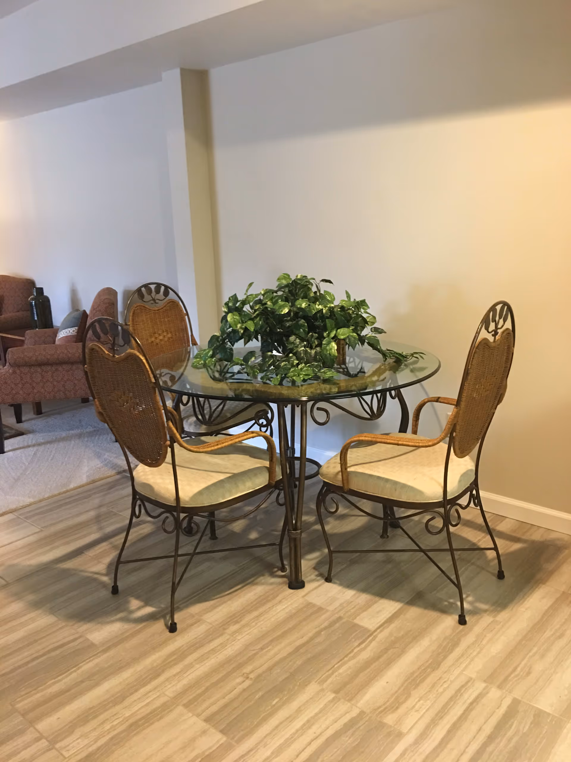 A small round glass-top table with a decorative metal base surrounded by four metal chairs with woven backs and cushioned seats. A green leafy plant centerpiece is on the table. In the background, there are upholstered armchairs and a side table with a decorative item, all set on a light-colored tiled floor and beige walls.