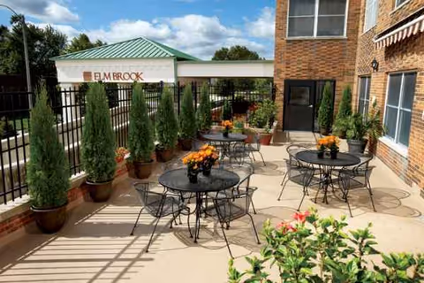 Outdoor terrace at Grove of Elmhurst with round tables, chairs, potted evergreens, and brick building.