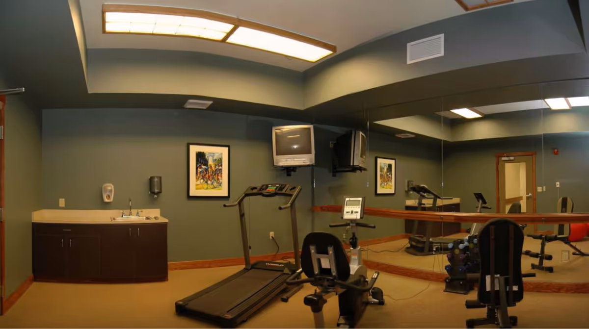 Small fitness room with a treadmill, recumbent bike, free weights, mirrored wall, sink cabinet and wall-mounted TV.