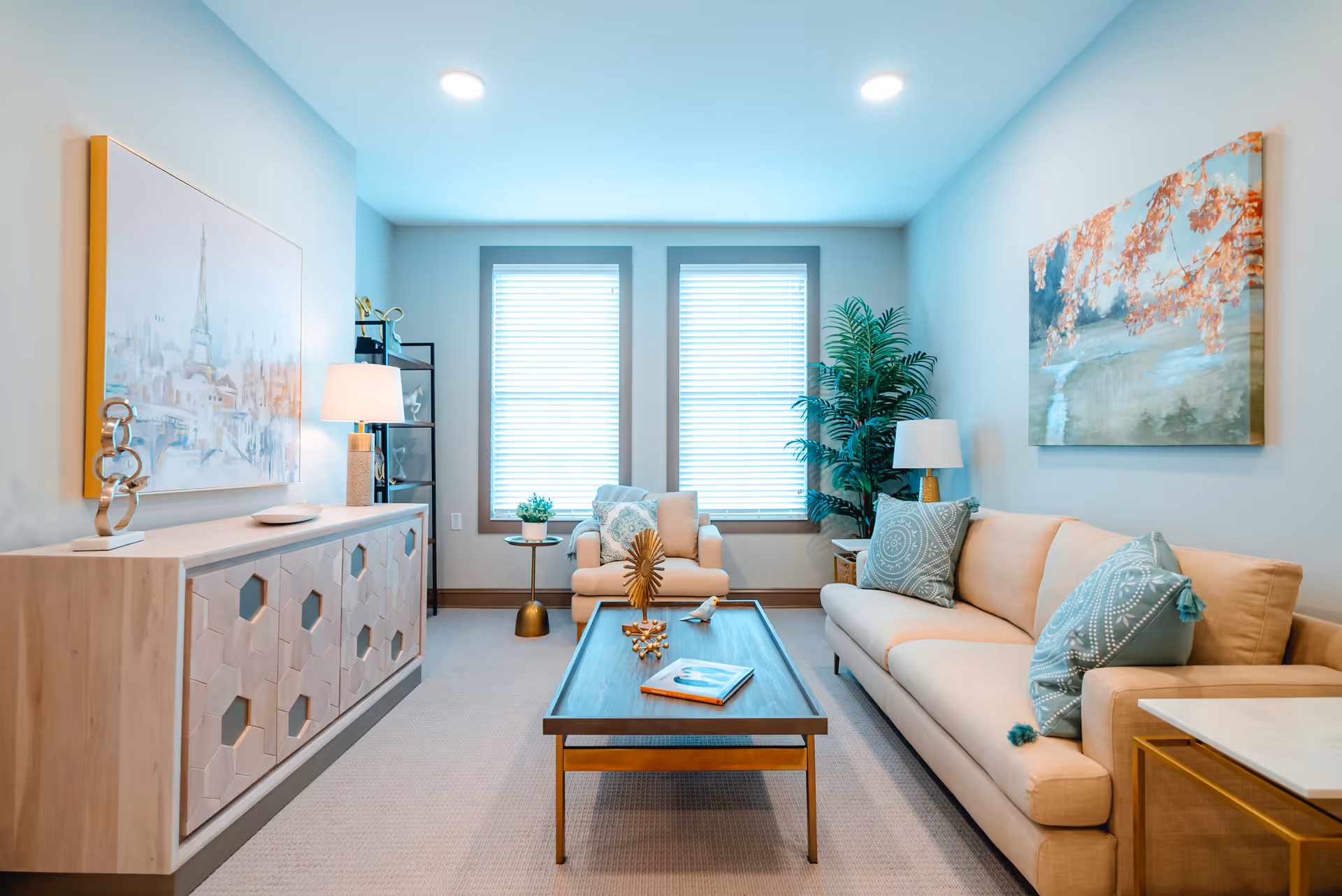 A bright and modern living room with a beige sofa adorned with blue patterned pillows, a matching armchair, a wooden coffee table with decorative items and a book, two large windows with blinds, a tall green plant in the corner, a side table with a lamp, a wooden sideboard with a lamp and decorative sculpture, and two large paintings on the walls.