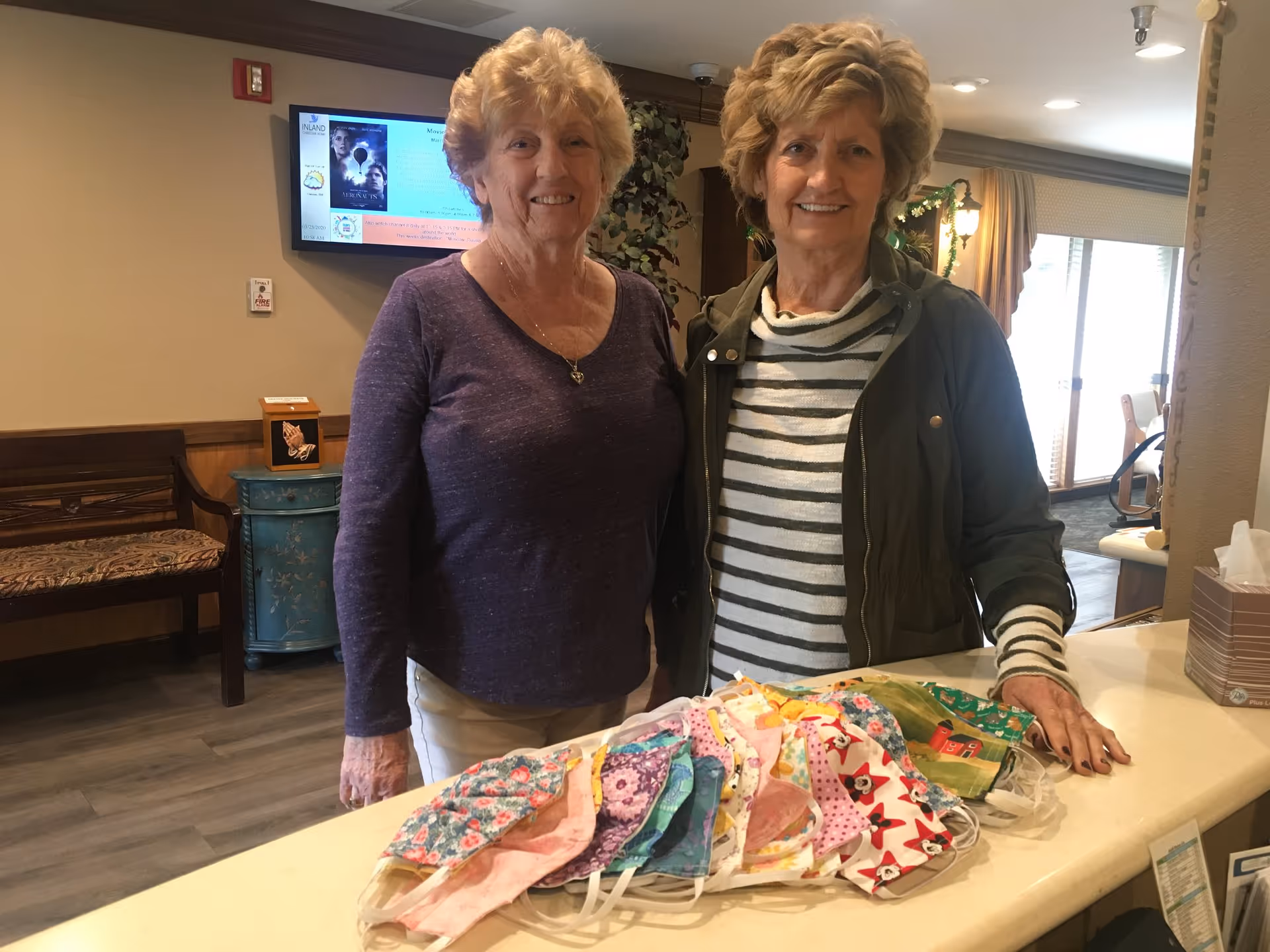 Two elderly women standing behind a counter displaying a variety of colorful face masks in a well-lit room with a bench, a small cabinet, and a television screen mounted on the wall in the background.