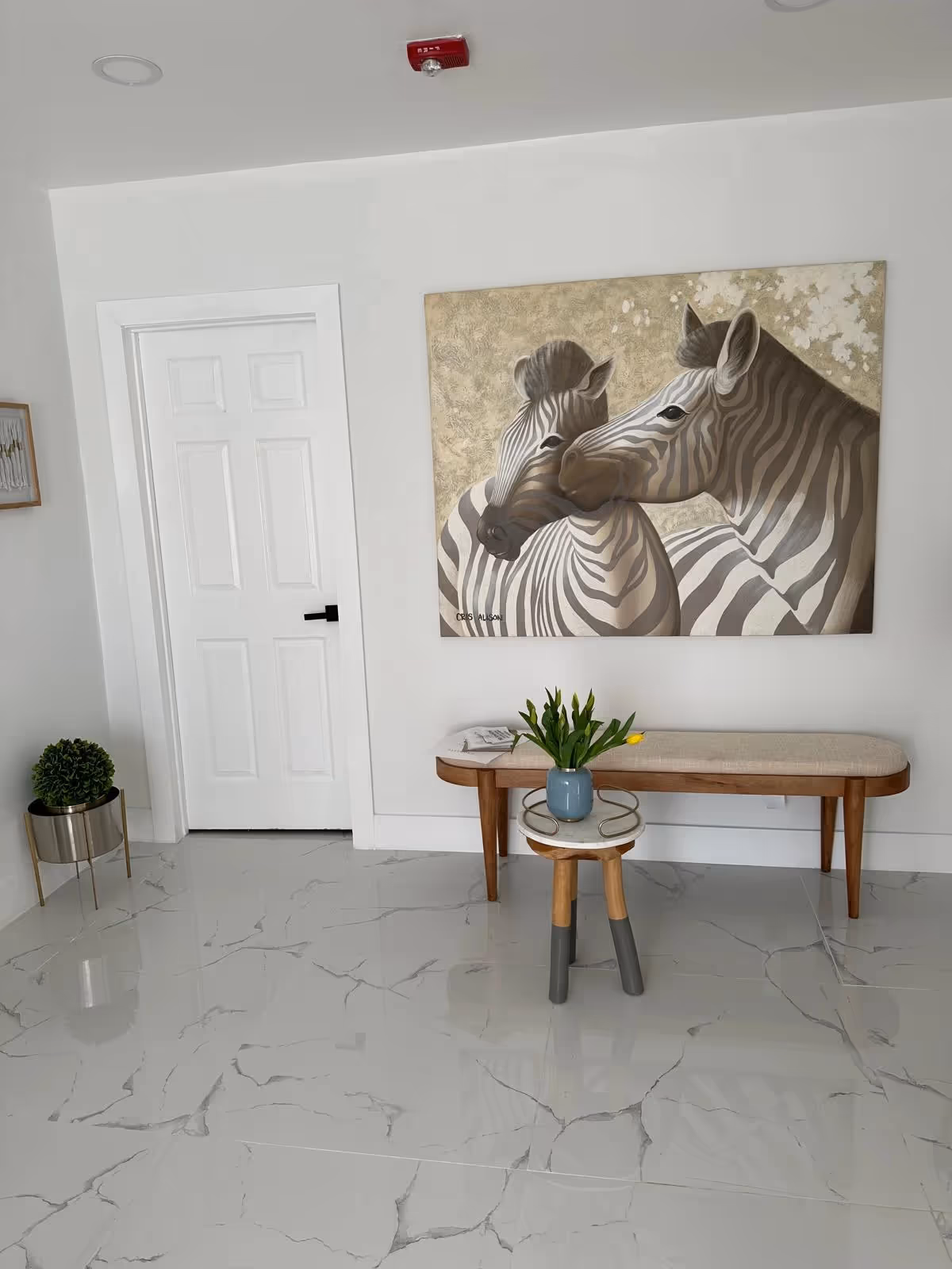 A bright, minimalist lobby area with a zebra painting, a wooden bench, a small round table with a vase, and a glossy white marble floor.
