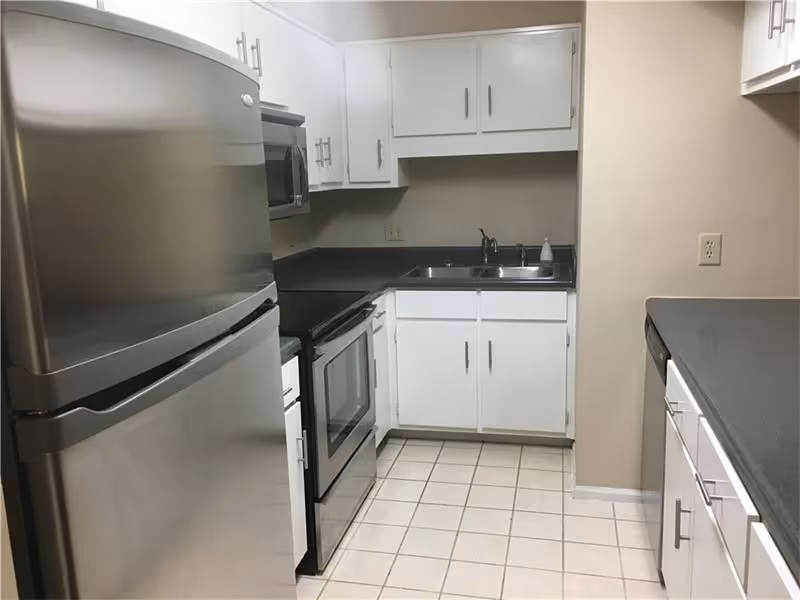 A compact kitchen with white cabinets, a stainless steel refrigerator, a microwave, an electric stove with oven, a double sink, and gray countertops. The floor is tiled with white square tiles.