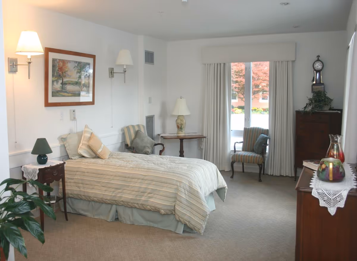 A cozy bedroom with a single bed covered in striped bedding, two striped armchairs with cushions, a wooden nightstand with a green lamp, a small wooden table with a decorative lamp, a wooden dresser with a clock and plants, and a large window with white curtains showing autumn foliage outside.