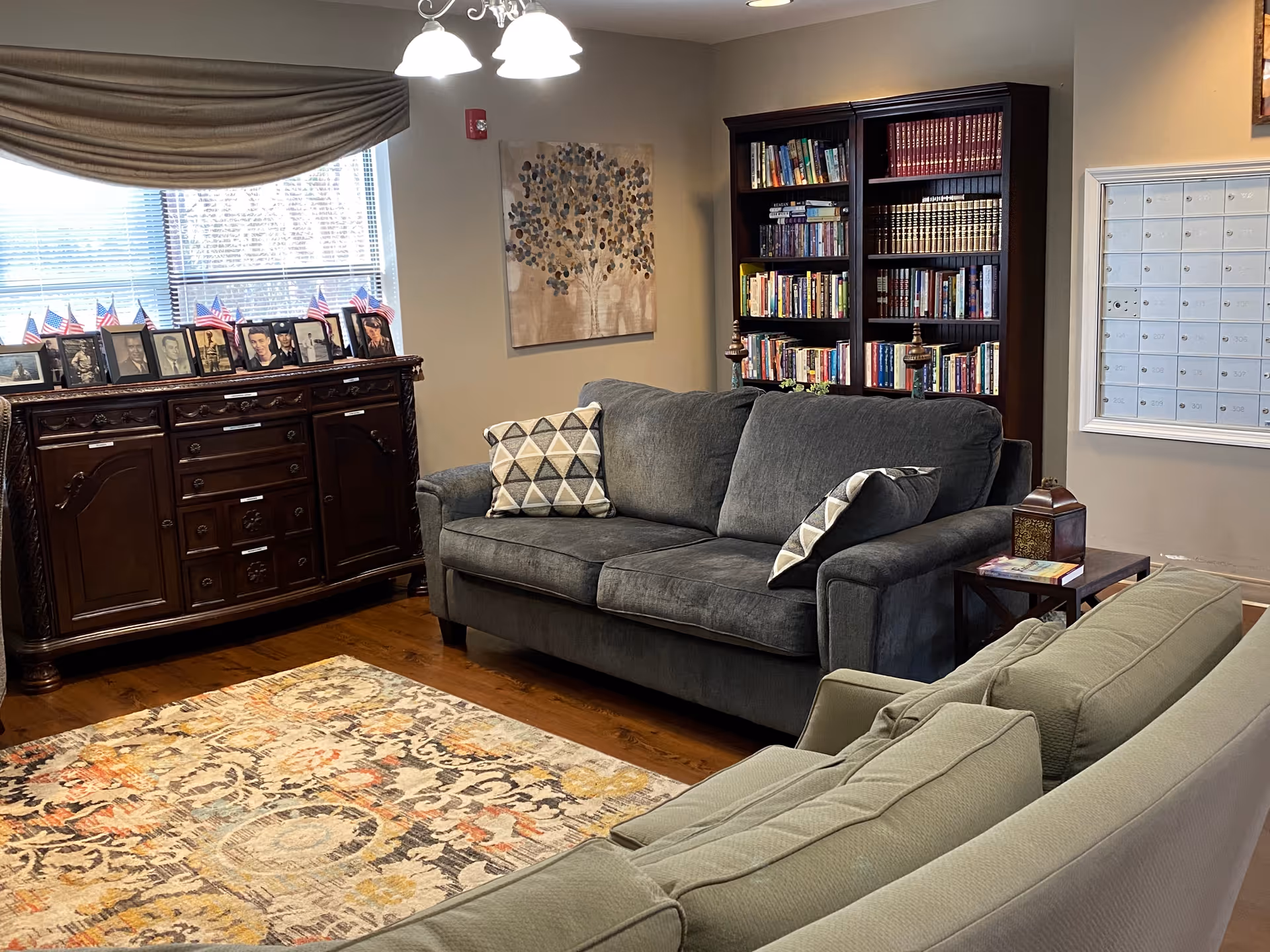 A cozy living room area with two sofas, one gray and one light green, arranged around a patterned rug on a wooden floor. There is a dark wooden bookshelf filled with books against the wall, and a wooden cabinet topped with framed photos and small American flags near a window with blinds and a brown valance. A painting of a tree hangs on the wall above the gray sofa, and a wall-mounted mailbox unit is visible on the right side.