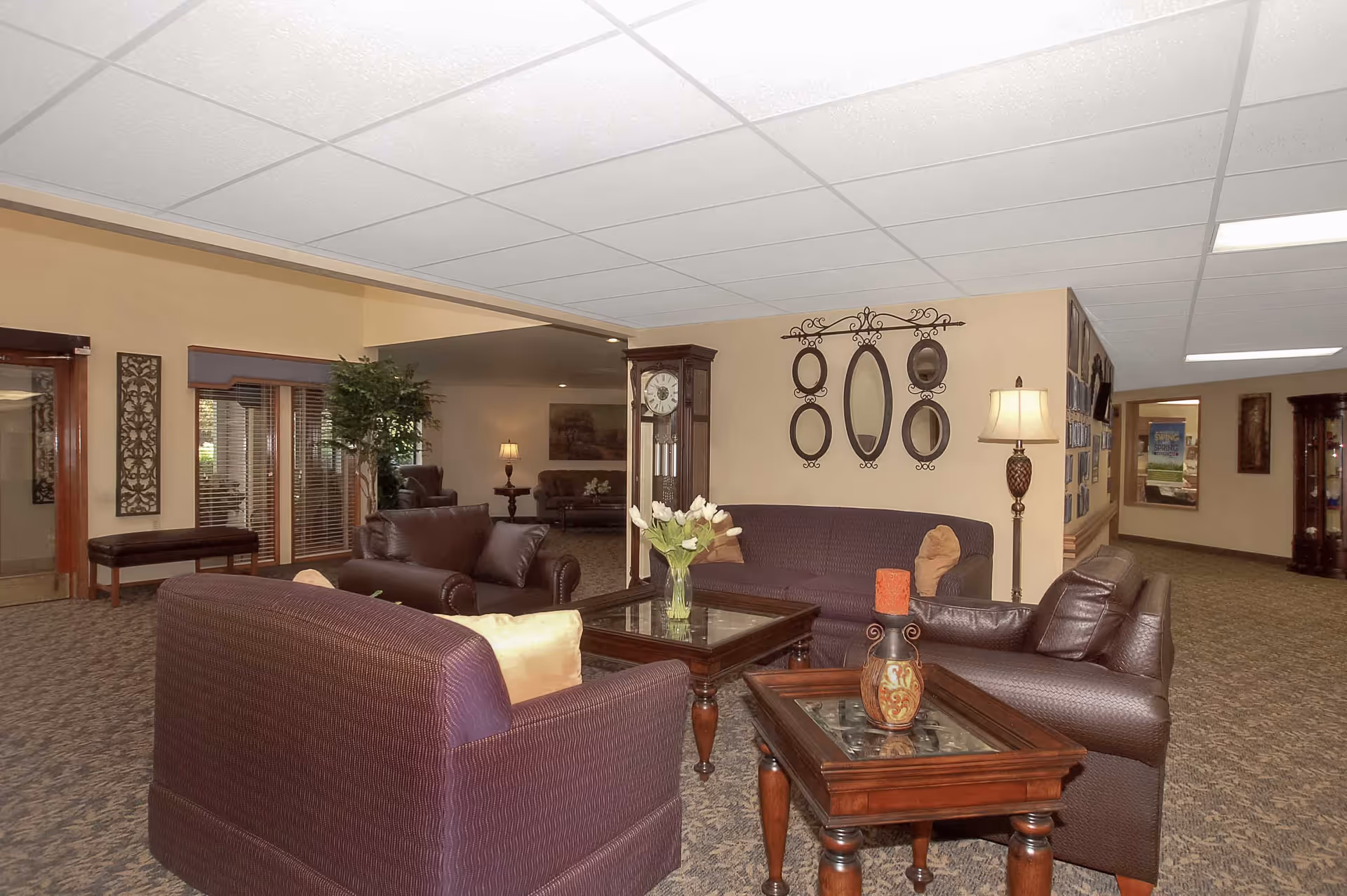 A cozy senior living facility lounge area with purple upholstered sofas and armchairs arranged around wooden coffee tables. The room features a grandfather clock, a floor lamp, decorative wall mirrors, and a vase with white tulips on the coffee table. The carpeted floor and beige walls create a warm atmosphere, with additional seating and lamps visible in the background.