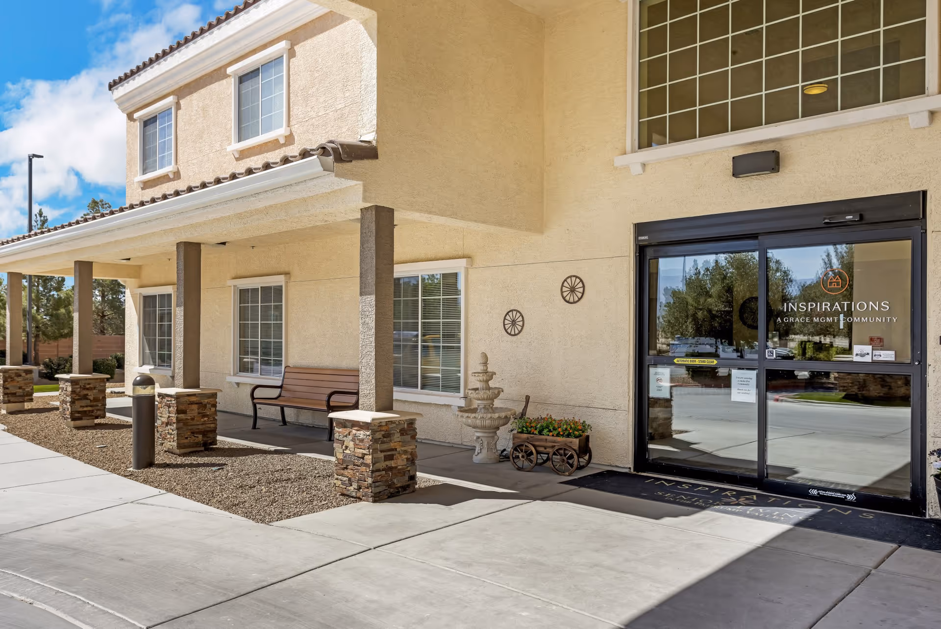 Exterior view of the entrance to Inspirations Of Pahrump facility showing a beige stucco building with large windows, a covered walkway supported by stone pillars, a bench, decorative wall hangings, a small fountain, and a flower cart near the glass sliding doors with the facility's name and logo.
