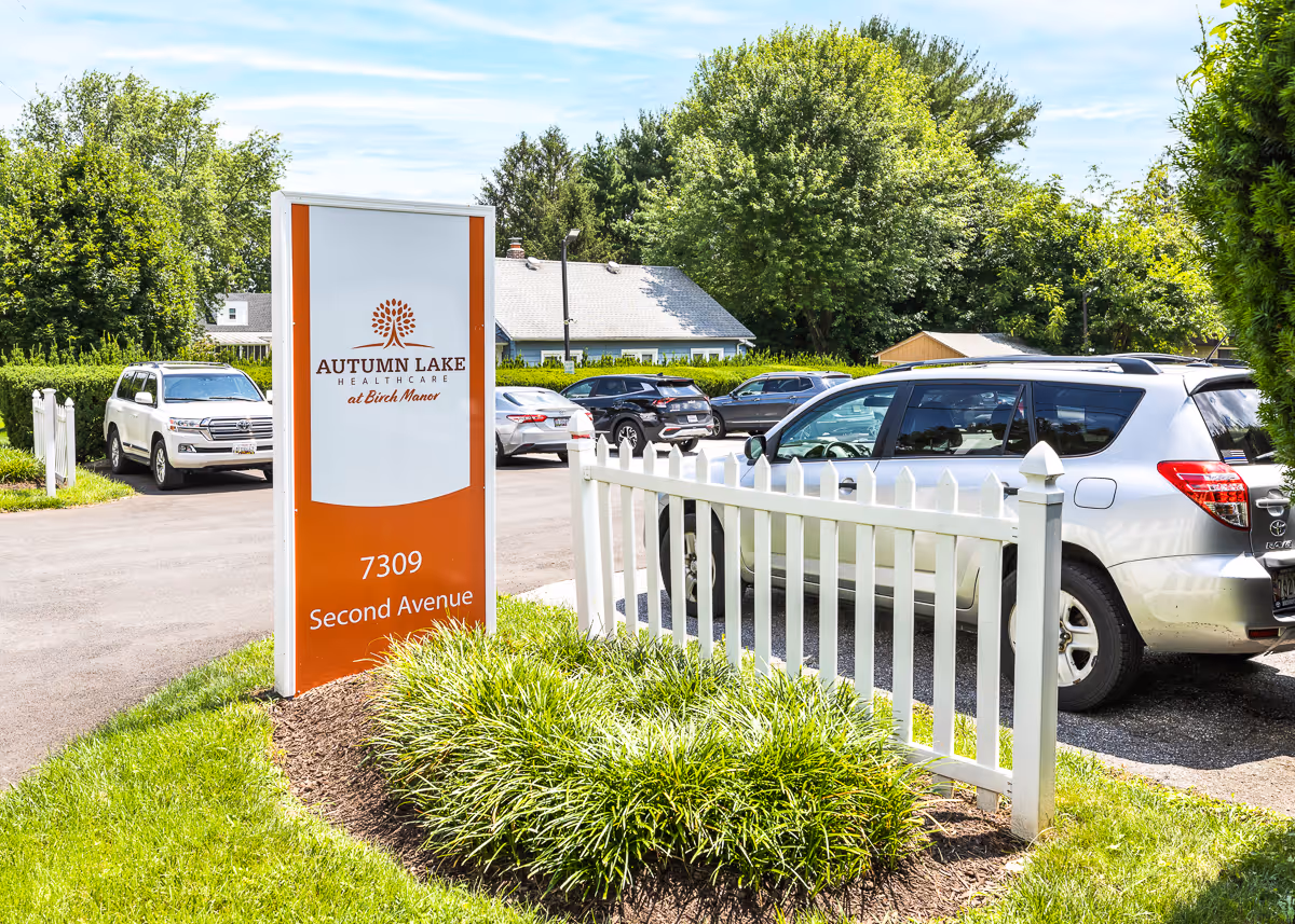 Outdoor view of the parking area at Autumn Lake Healthcare at Birch Manor, showing a white picket fence, green bushes, several parked cars, and a tall sign with the facility's name and address 7309 Second Avenue.