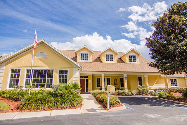Exterior view of a single-story assisted living facility building with a beige facade, multiple windows, a front porch with columns, an American flag on a flagpole, landscaped bushes and plants, a parking area, and a tree on the right side under a partly cloudy blue sky.