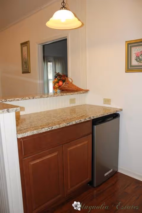 A small kitchen area with a granite countertop, wooden cabinets below, and a compact stainless steel refrigerator. A decorative basket with artificial fruit is placed on the counter. The wall has two framed floral pictures, and a hanging light fixture is above the counter.
