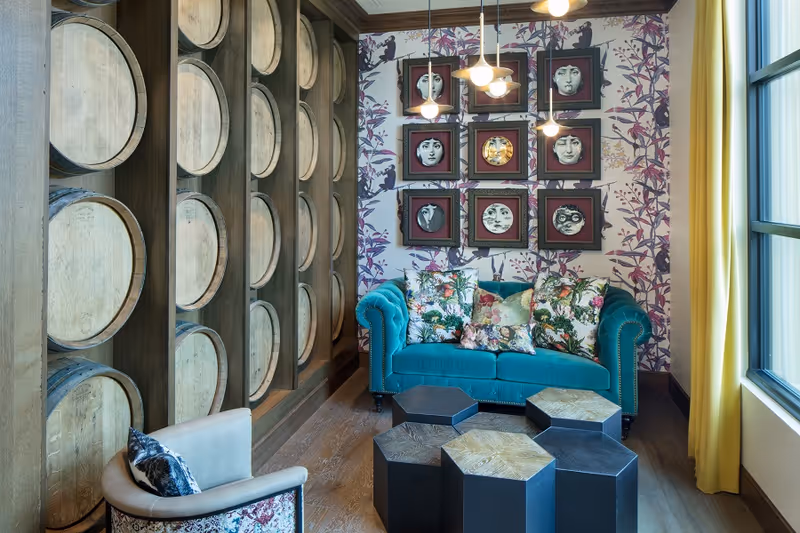A cozy living room area with a teal velvet sofa adorned with floral patterned pillows. In front of the sofa are five hexagonal wooden tables in varying shades. To the left, a wall features a decorative display of wooden barrels arranged vertically in cubby-like shelves. The back wall has wallpaper with a purple floral and bird pattern and is decorated with nine framed circular artworks depicting various faces. A large window with yellow curtains allows natural light to fill the room.