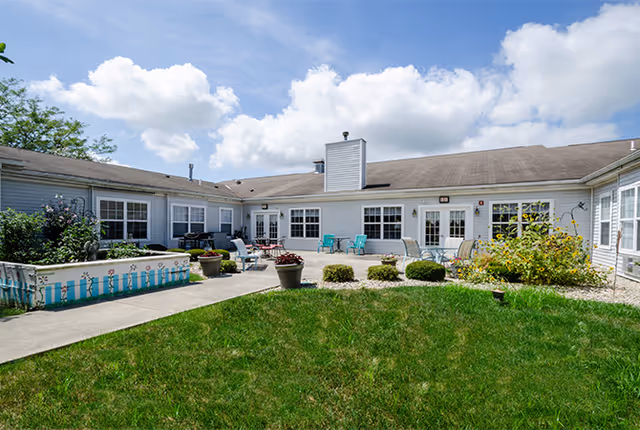 Outdoor courtyard area of a senior living facility with green grass, potted plants, garden beds, and patio furniture under a partly cloudy sky.