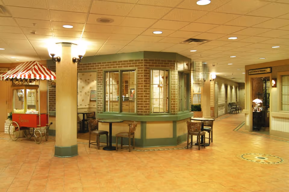 Indoor common area with a popcorn machine on wheels with a red and white striped canopy, a central brick and green counter area with windows, several small tables with chairs, and a hallway leading to other rooms. The floor is tiled and the ceiling has recessed lighting.