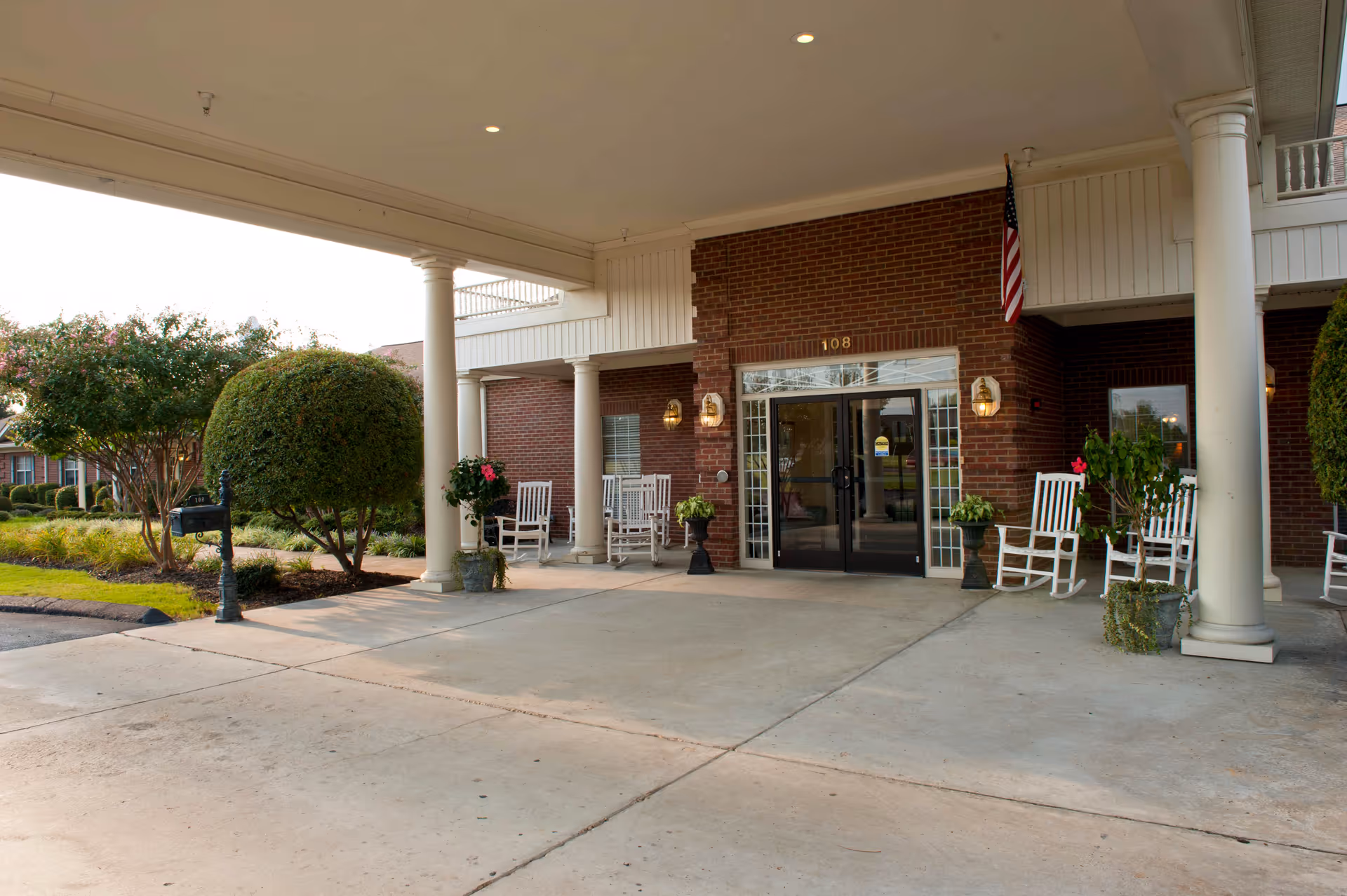 Covered entrance area of a brick building with white columns and several white rocking chairs. There are potted plants near the entrance and an American flag mounted on the wall. The entrance has glass double doors with the number 108 above them. Well-maintained landscaping with trimmed bushes and trees is visible to the left.
