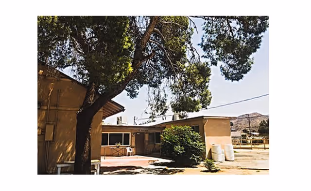 Exterior view of a single-story building with a tree in the foreground and some bushes near the building. The sky is clear and there are hills visible in the background.