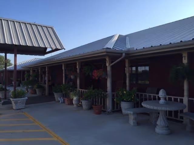 Covered front entrance of a brick building with a metal roof, potted plants, hanging baskets, and stone benches on a concrete driveway.