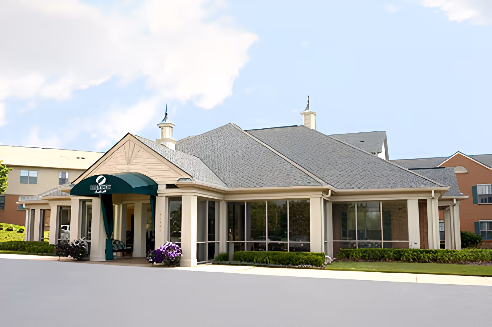 Exterior view of a senior living facility building with a peaked roof, large windows, and a green awning over the entrance. The building is surrounded by neatly trimmed bushes and flowers, with a clear sky in the background.
