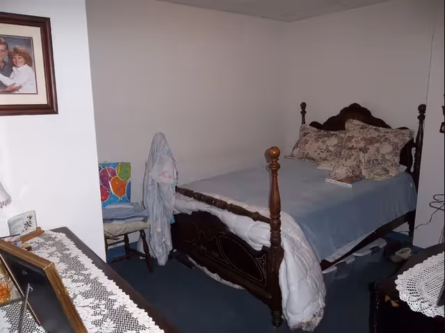 A small bedroom with a wooden bed frame, a bed made with a blue blanket and floral pillows, a chair with a colorful cushion and a piece of clothing draped over it, a dresser with a lace runner, and a framed photo on the wall.