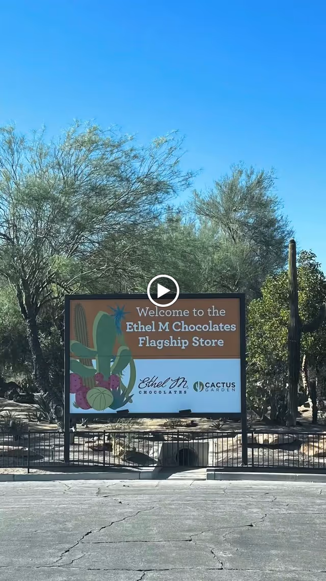 Outdoor scene showing a sign that reads 'Welcome to the Ethel M Chocolates Flagship Store' with illustrations of cacti and desert plants in the background under a clear blue sky.