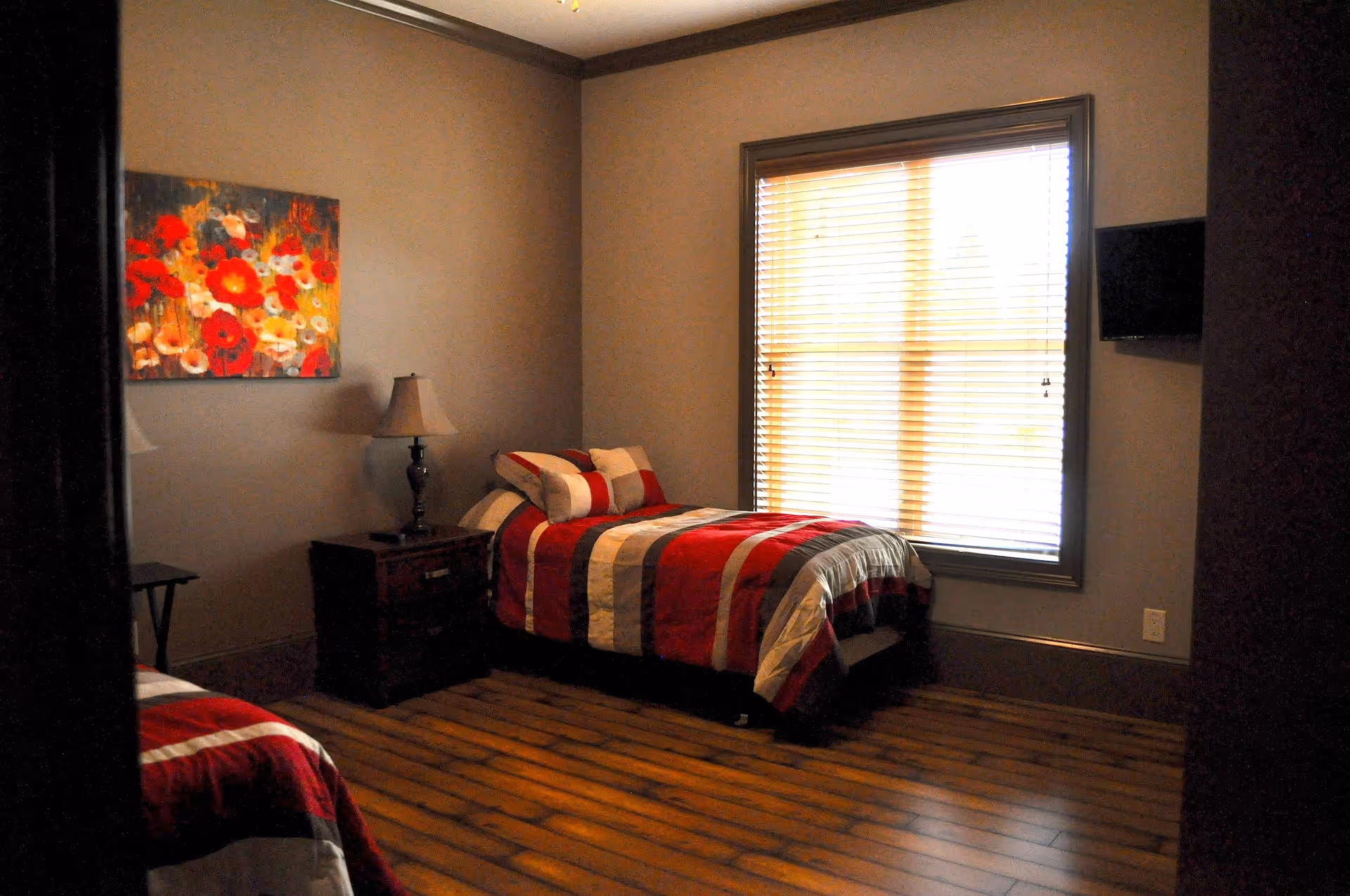 A bedroom with two twin beds featuring red, white, and gray striped bedding. There is a wooden nightstand with a lamp between the beds, a colorful floral painting on the wall, a window with blinds, and a wall-mounted TV.