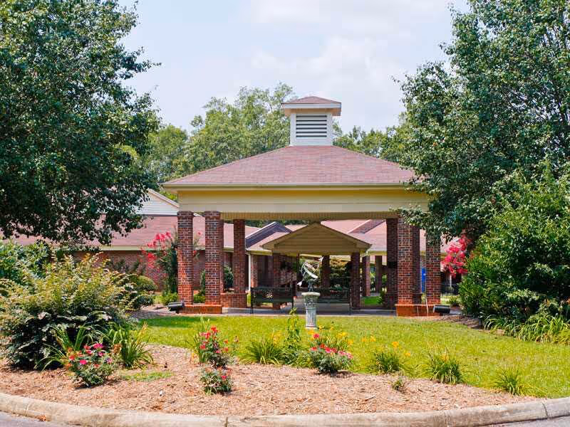 Outdoor view of a covered entrance area with brick pillars and a red roof at Woodbridge of Clinton, surrounded by green trees, shrubs, and colorful flowers under a partly cloudy sky.