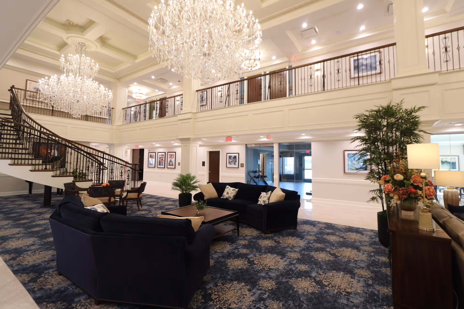 Spacious and elegant living room area in a senior living facility with two large chandeliers hanging from a high ceiling. The room features a grand staircase with ornate railings, two dark blue sofas with decorative pillows, a coffee table, and several plants. The walls are adorned with framed artwork, and there is a second-floor balcony overlooking the area.