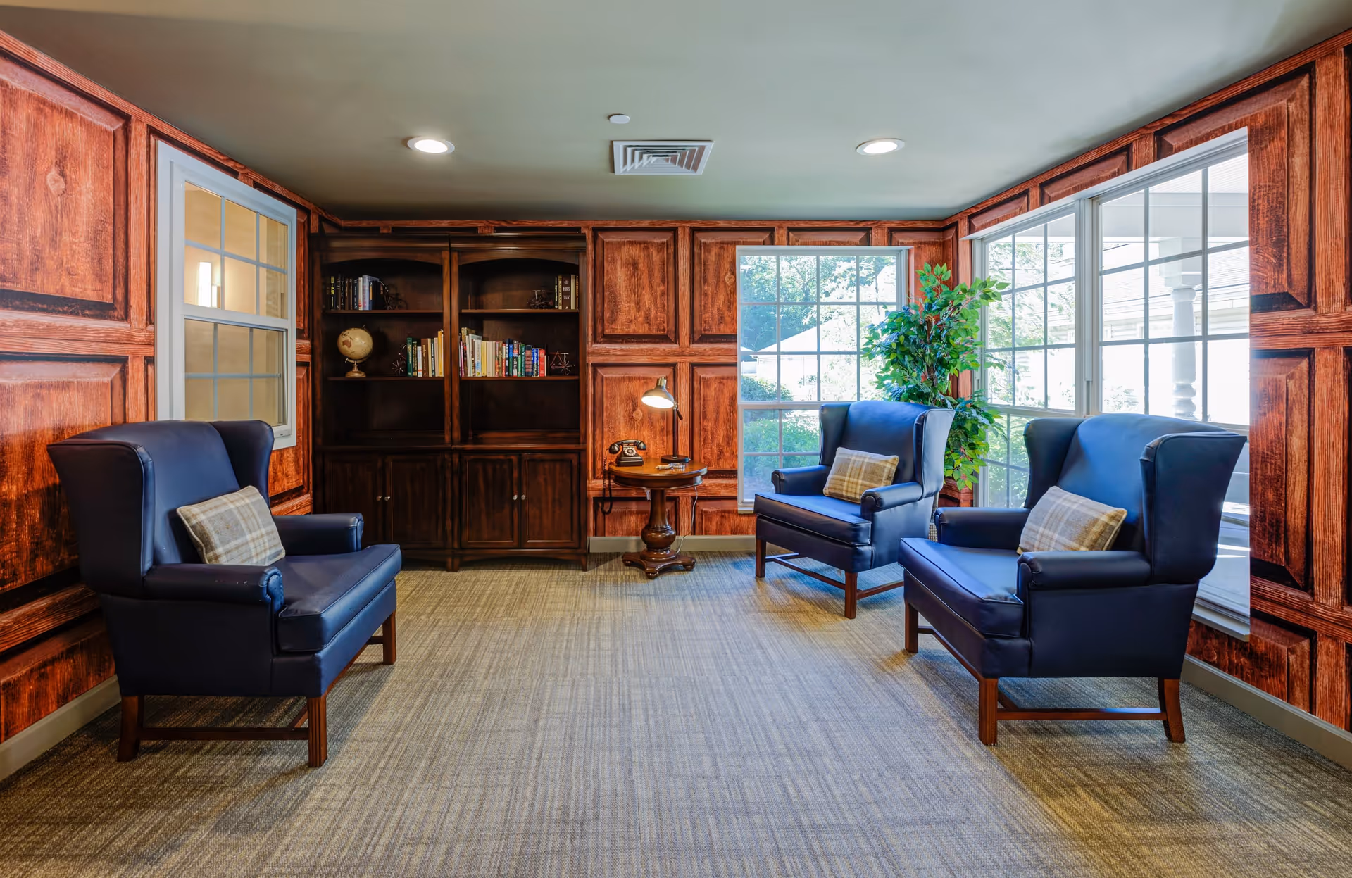 A cozy sitting area with three dark blue armchairs, each with a plaid cushion, arranged around a small wooden side table with a vintage telephone and a lamp. The room has wood-paneled walls, a built-in bookshelf with books and a globe, large windows letting in natural light, and a potted plant near the window.