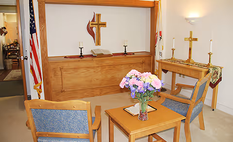 A small chapel or prayer room with two wooden chairs with blue upholstery facing a wooden altar. On the altar, there is an open Bible, two candles, and a wooden cross mounted on the wall above it. A small table with a vase of colorful flowers and a closed book is placed between the chairs. An American flag and another flag stand in the corners of the room. Another small table with two lit candles and a gold cross is visible to the right.