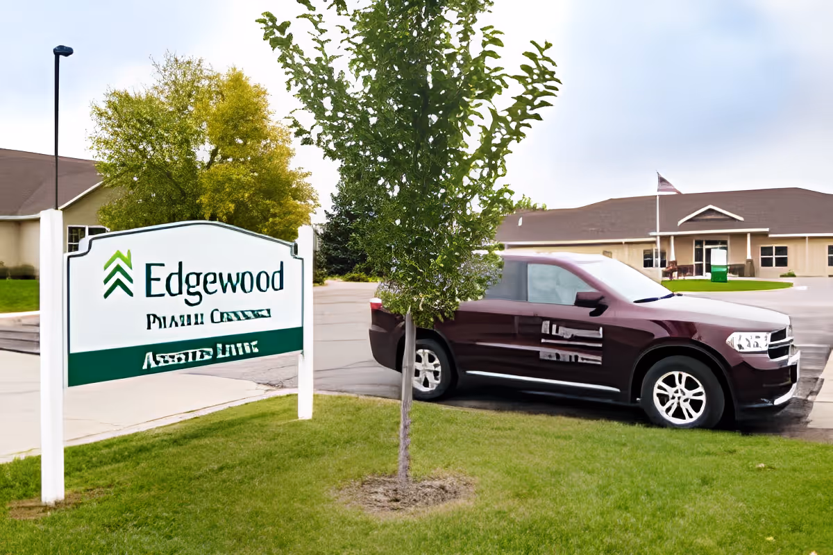 Outdoor view of Edgewood Prairie Crossings Assisted Living facility showing a white sign with the facility name, a small tree in the foreground, a parked maroon SUV, and a building with an American flag in the background.