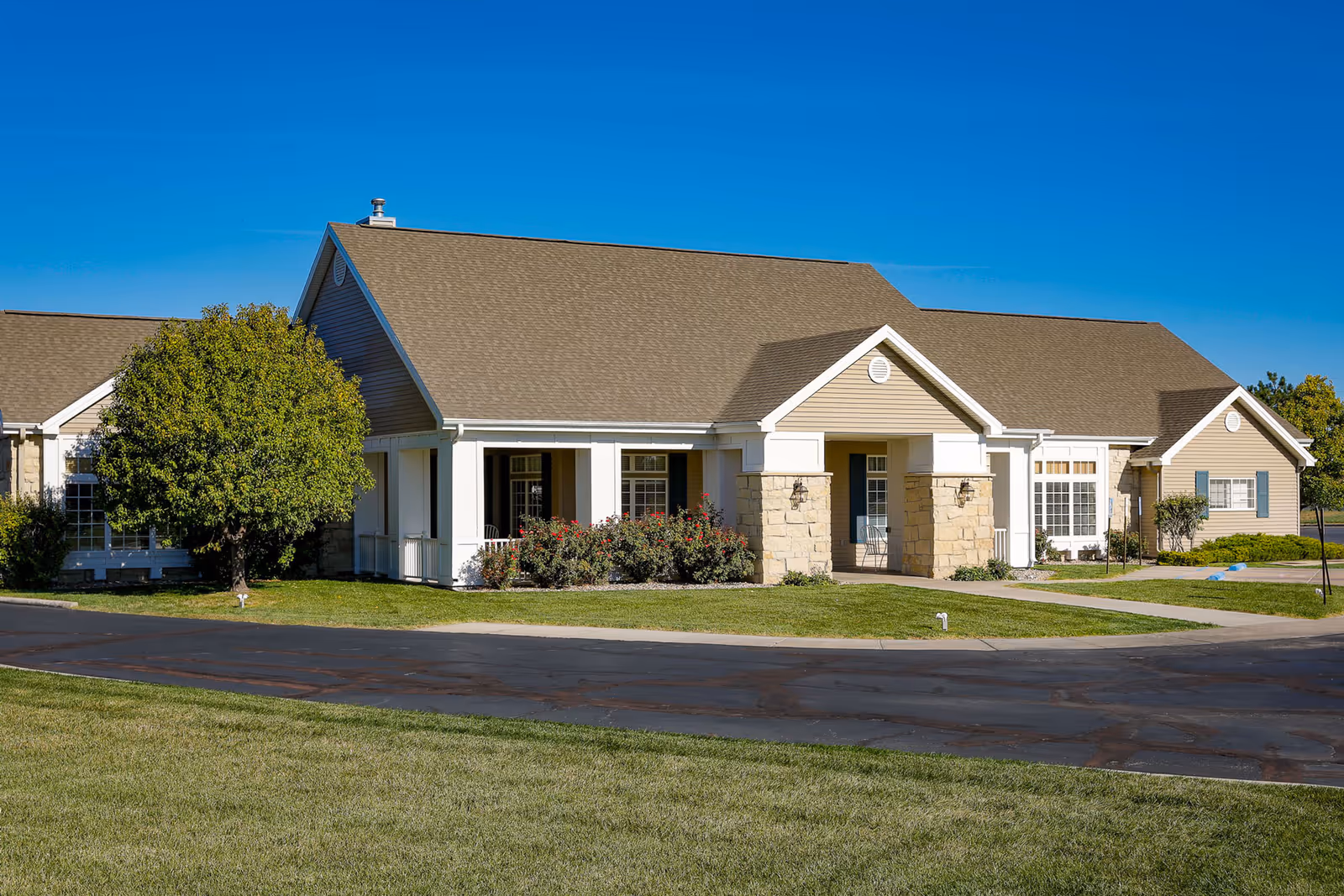 Exterior view of a single-story assisted living facility building with beige siding, stone accents, a gabled roof, and a small covered porch. The building is surrounded by a well-maintained lawn, a tree, and shrubs under a clear blue sky.