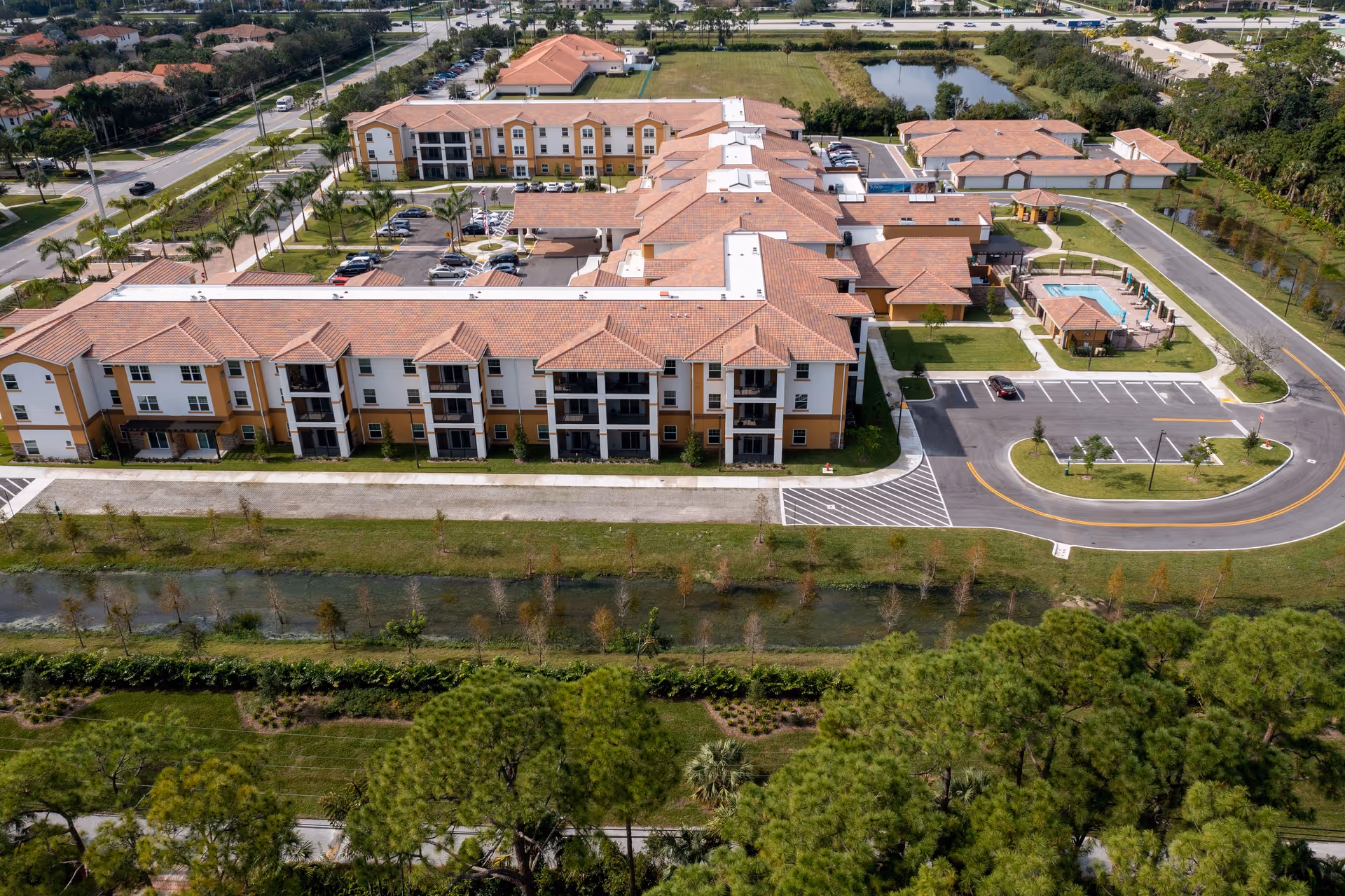 Aerial view of Palomino Gardens Retirement Community showing multiple connected buildings with terracotta roofs, parking lots, a swimming pool area, surrounding roads, and landscaped greenery including trees and a water canal.
